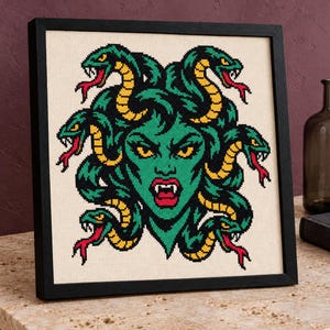 May include: A framed cross-stitch embroidery featuring a green-skinned Medusa figure with yellow and green snakes for hair, yellow eyes, and red lips, set against a cream-colored fabric background within a simple black square frame.