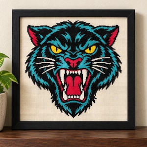 May include: A framed cross-stitch embroidery featuring the head of a snarling panther. The design uses black, teal, and red thread on a cream-colored fabric background. The panther has bright yellow eyes, white fangs, and a red mouth.