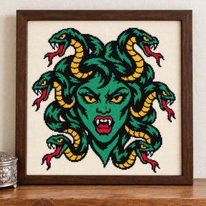 May include: A framed cross-stitch embroidery featuring a green-skinned figure with yellow eyes and fangs, surrounded by multiple green and yellow snakes with red tongues, all set against a cream-colored fabric background.