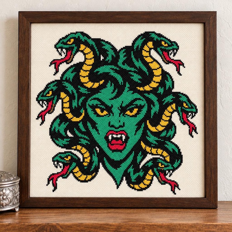 May include: A framed cross-stitch embroidery featuring a green-skinned figure with yellow eyes and fangs, surrounded by multiple green and yellow snakes with red tongues, all set against a cream-colored fabric background.