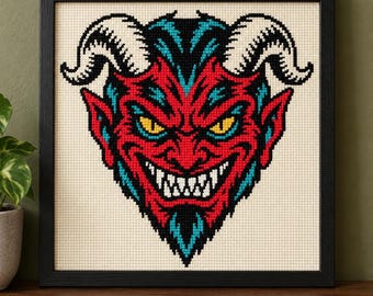 May include: A framed cross-stitch embroidery featuring a stylized devil face with red skin, bright yellow eyes, sharp white teeth, and large white horns. The design uses black and teal accents against a cream-colored fabric background.