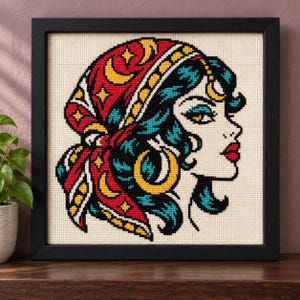 May include: A framed cross-stitch embroidery featuring a profile view of a person with teal hair wearing a red headscarf decorated with yellow crescent moons and stars. The person has a gold hoop earring and bright red lips on a cream fabric.