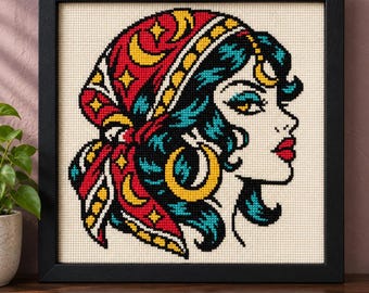 May include: A framed cross-stitch embroidery featuring a profile view of a person with teal hair wearing a red headscarf decorated with yellow crescent moons and stars. The person has a gold hoop earring and bright red lips on a cream fabric.