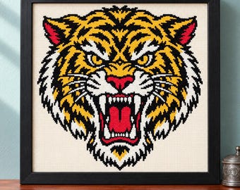 May include: A framed cross-stitch embroidery featuring a stylized tiger head with an open mouth, displaying sharp teeth and a red tongue. The tiger is rendered in vibrant yellow, black, and white thread against a cream-colored fabric background.