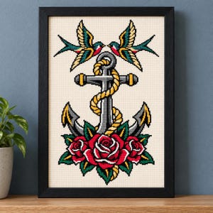 May include: A framed cross-stitch embroidery featuring a central gray anchor wrapped in a yellow rope, flanked by two swallows at the top and three red roses with green leaves at the base, all set against a cream-colored fabric background.