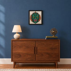 May include: A framed cross-stitch embroidery featuring a green, scaly creature with yellow eyes, sharp teeth, and orange fins, displayed on a wall above a wooden cabinet with a table lamp and a small metal globe on top.