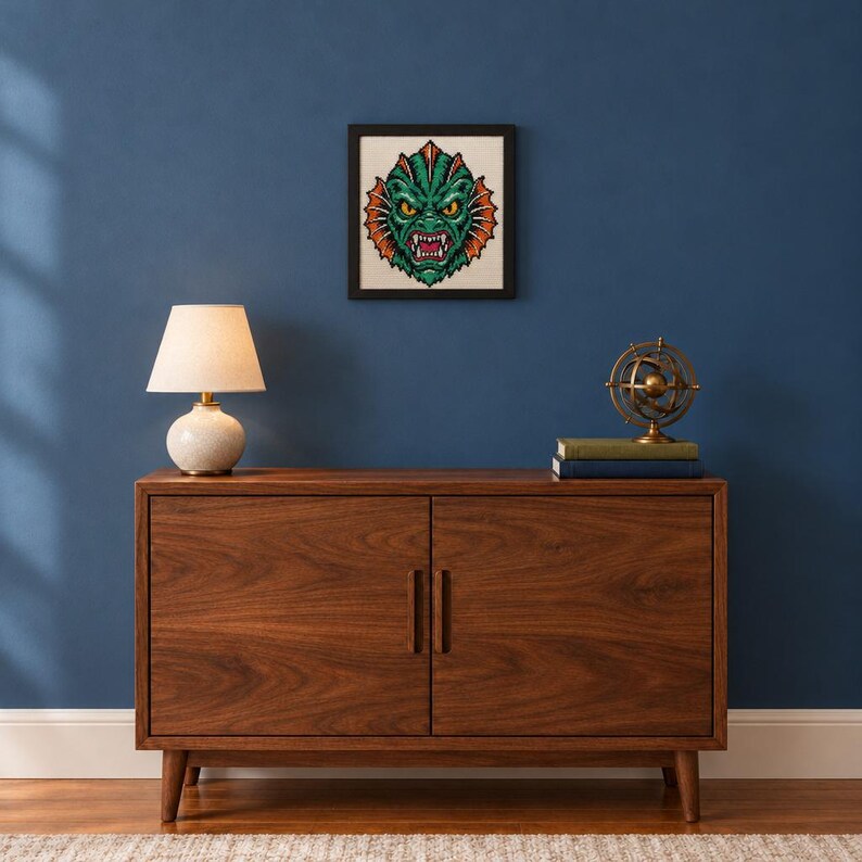 May include: A framed cross-stitch embroidery featuring a green, scaly creature with yellow eyes, sharp teeth, and orange fins, displayed on a wall above a wooden cabinet with a table lamp and a small metal globe on top.