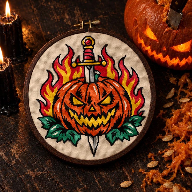 May include: A circular embroidery hoop containing a cross-stitch pattern of an orange jack-o'-lantern pierced by a silver dagger, surrounded by yellow and red flames and green leaves, set against a cream-colored fabric background.