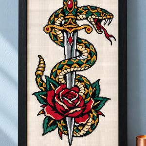 Può includere: Un ricamo a punto croce incorniciato raffigurante un pugnale argentato che trafigge una rosa rossa, con un serpente dal motivo verde e giallo avvolto attorno alla lama. Il disegno è su un tessuto color crema all'interno di una cornice nera.