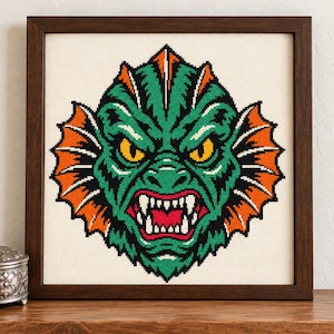 May include: A framed cross-stitch embroidery featuring a stylized green creature head with sharp white teeth, yellow eyes, and vibrant orange fins, set against a cream-colored fabric background within a dark brown wooden frame.