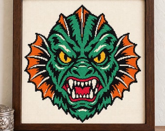 May include: A framed cross-stitch embroidery featuring a stylized green creature head with sharp white teeth, yellow eyes, and vibrant orange fins, set against a cream-colored fabric background within a dark brown wooden frame.