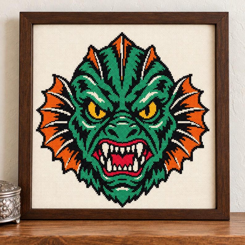 May include: A framed cross-stitch embroidery featuring a stylized green creature head with sharp white teeth, yellow eyes, and vibrant orange fins, set against a cream-colored fabric background within a dark brown wooden frame.