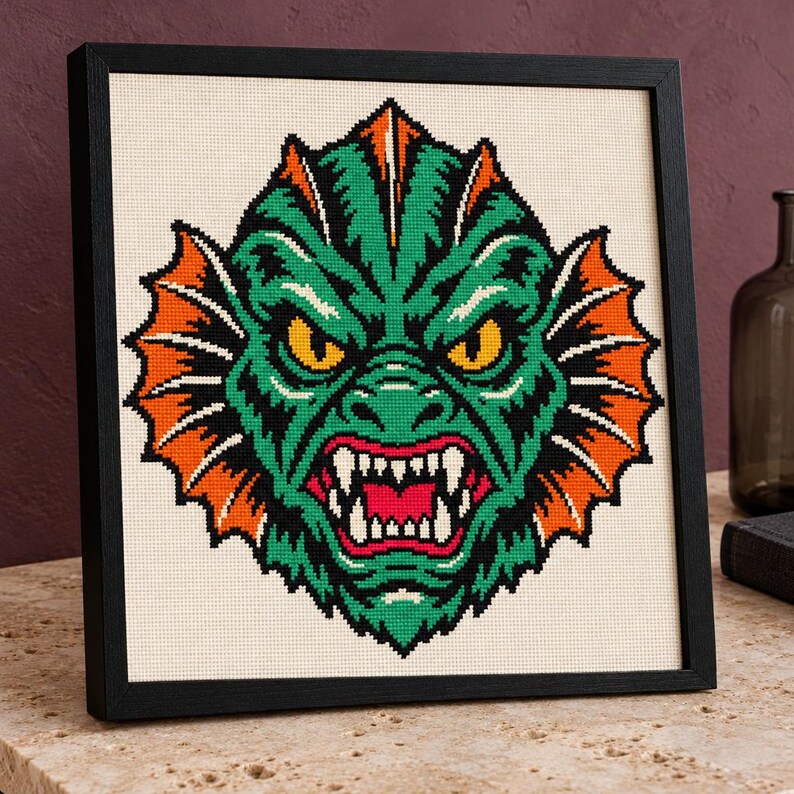 May include: A framed cross-stitch embroidery featuring a stylized green creature head with orange fins, yellow eyes, and a wide-open mouth showing sharp white teeth. The design is centered on a cream-colored fabric background within a black frame.