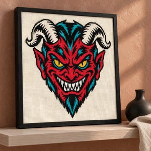 Grinning devil cross stitch in a black frame on a shelf at an angle