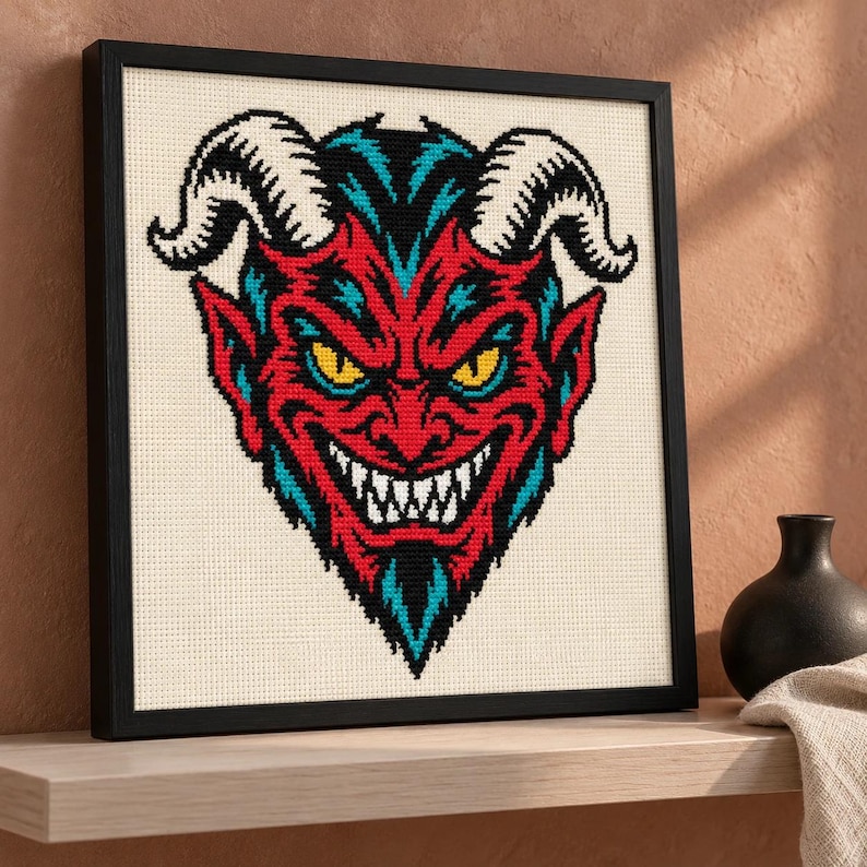 Grinning devil cross stitch in a black frame on a shelf at an angle