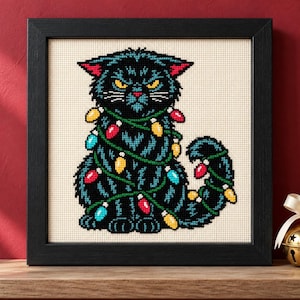 May include: A framed cross-stitch embroidery featuring a black cat with yellow eyes and pink inner ears, wrapped in a string of colorful red, yellow, and blue Christmas lights. The cat has a grumpy expression and is set against a cream fabric background.