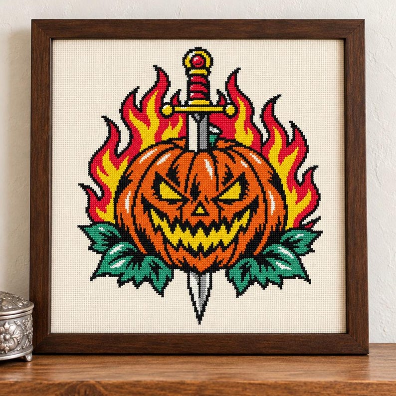 May include: A cross-stitch embroidery in a dark wood frame featuring a jack-o'-lantern with a jagged grin, pierced by a silver sword with a red and gold hilt. The pumpkin is surrounded by bright yellow and red flames and green leaves.