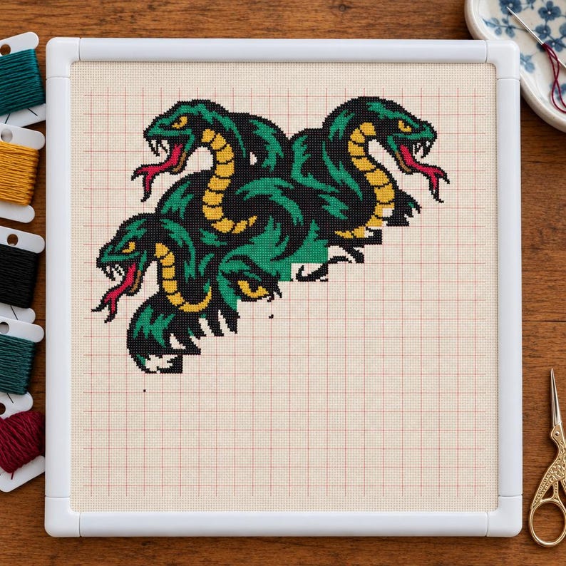 May include: A cross-stitch project in a white plastic frame featuring a multi-headed snake design. The snakes are stitched in shades of dark green, black, and yellow with red tongues and white fangs, set against a cream-colored grid fabric.