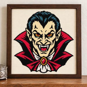 May include: A cross-stitch embroidery of a vampire with dark hair, pale skin, and yellow eyes, wearing a high-collared black cape with a red lining and a red gemstone brooch, displayed in a square dark brown wooden frame.