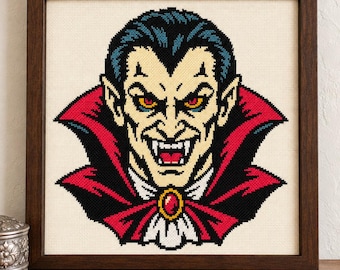 May include: A cross-stitch embroidery of a vampire with dark hair, pale skin, and yellow eyes, wearing a high-collared black cape with a red lining and a red gemstone brooch, displayed in a square dark brown wooden frame.