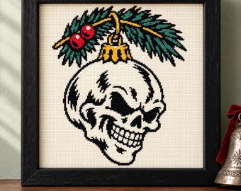 May include: A framed cross-stitch embroidery featuring a white skull shaped like a Christmas ornament, topped with a gold cap, green pine needles, and two red berries, all set against a cream-colored fabric background.