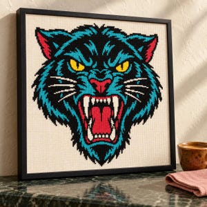 Snarling panther cross stitch in black frame leaning on a wood shelf