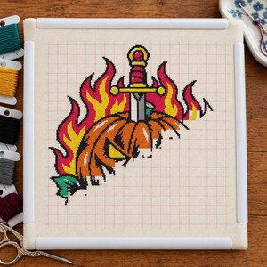May include: A cross-stitch project on white fabric in a square frame featuring an orange jack-o'-lantern pierced by a silver sword with a gold hilt, surrounded by stylized yellow and red flames, with embroidery floss spools nearby.