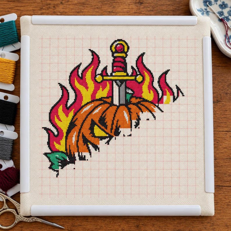 May include: A cross-stitch project on white fabric in a square frame featuring an orange jack-o'-lantern pierced by a silver sword with a gold hilt, surrounded by stylized yellow and red flames, with embroidery floss spools nearby.