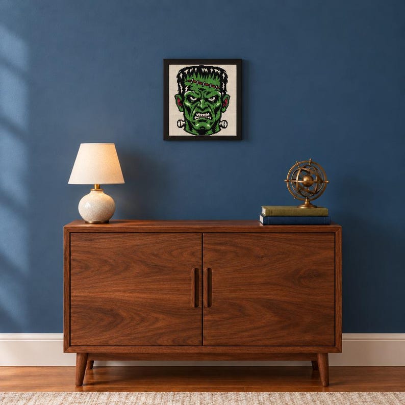 Puede incluir: Una pieza de arte mural cuadrada enmarcada que presenta una ilustraci&oacute;n pixelada de un monstruo de piel verde con cabello oscuro, frente cosida y pernos de metal en el cuello, exhibida en un marco negro sobre una pared azul oscuro.