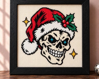 May include: A cross-stitch embroidery in a black frame featuring a skull wearing a red and white Santa hat with green holly leaves and red berries. The skull has blue star-shaped eyes and is surrounded by two small yellow four-pointed stars.