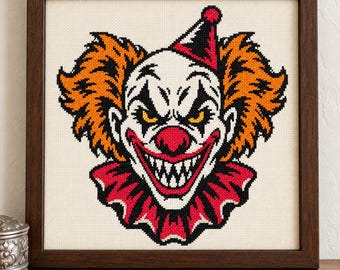 May include: A framed cross-stitch embroidery featuring a clown face with bright orange hair, a red nose, and a wide, sharp-toothed grin. The clown wears a red and black pointed hat and a ruffled collar, all set against a cream-colored fabric background.