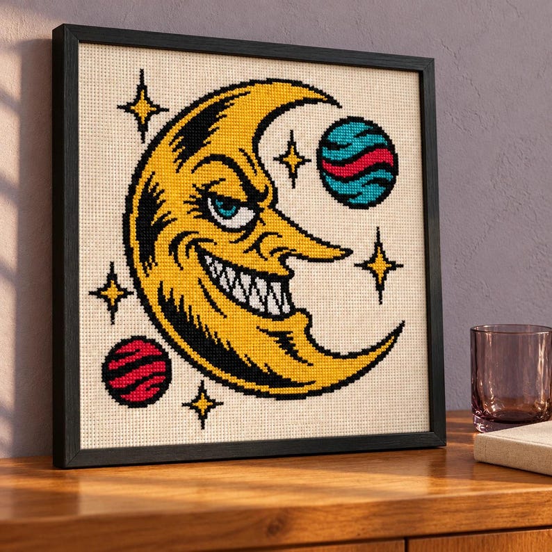 Crescent moon cross stitch in a black frame on a wood shelf with an amber bottle