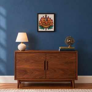 May include: A square framed cross-stitch art piece featuring a jack-o'-lantern pierced by a sword, surrounded by orange flames and green leaves. The artwork is displayed on a dark blue wall above a wooden cabinet with a lamp and books.