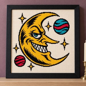May include: A framed cross-stitch embroidery featuring a yellow crescent moon with a mischievous face, sharp teeth, and blue eyes. The design includes two small planets with red and blue stripes, surrounded by four yellow four-pointed stars.