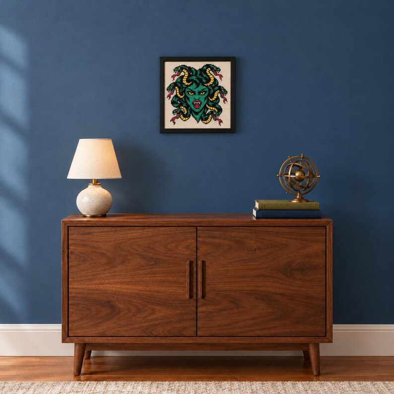 May include: A dark wood sideboard with two doors and vertical handles. On top sits a beige lamp with a round base and a brass-colored armillary sphere on two books. A framed square print of a green Medusa with snakes hangs on the blue wall.