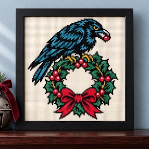 Straight-on view of the finished Raven Christmas Wreath Cross Stitch in a black frame on a wooden shelf