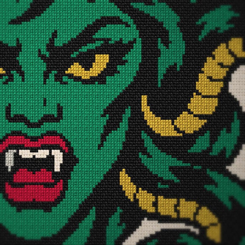 May include: A cross-stitch embroidery piece featuring a green-skinned character with yellow horns, sharp white fangs, and bright red lips. The design is set against a black background, with the character's face rendered in detailed green stitches.