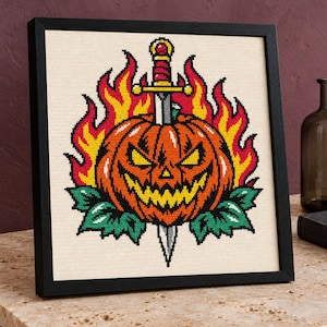 May include: A framed cross-stitch embroidery featuring a jack-o'-lantern pierced by a silver dagger. The pumpkin is orange with yellow eyes and a jagged mouth, surrounded by bright yellow and red flames and green leaves on a cream fabric.