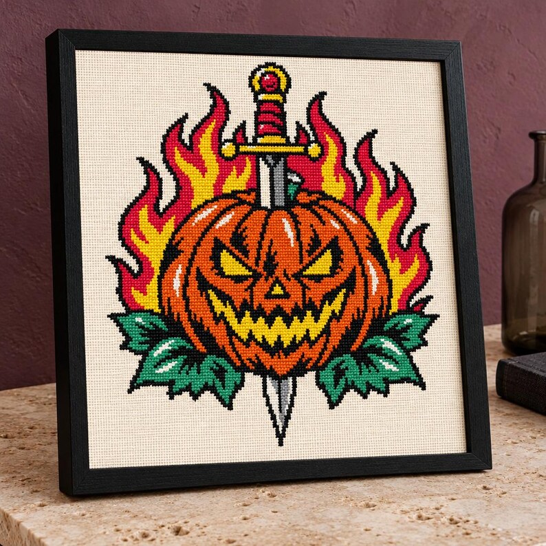 May include: A framed cross-stitch embroidery featuring a jack-o'-lantern pierced by a silver dagger. The pumpkin is orange with yellow eyes and a jagged mouth, surrounded by bright yellow and red flames and green leaves on a cream fabric.