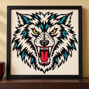 May include: A square framed cross-stitch embroidery featuring a stylized wolf head with white, black, and teal fur, piercing yellow eyes, and an open mouth revealing sharp teeth and a bright red tongue, set against a cream-colored fabric background.