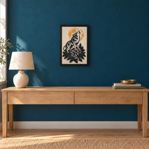 May include: A framed wall art print featuring a grey wolf howling at a large yellow moon, perched on a rocky outcrop surrounded by green pine trees. The illustration is rendered in a graphic style with bold lines and a cream-coloured background.