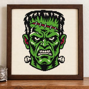 May include: A framed cross-stitch embroidery featuring a green-skinned monster with black hair, a stitched forehead scar, and metallic neck bolts. The design uses black, white, and red thread on a cream-colored fabric, enclosed in a dark wood frame.