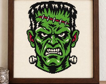 May include: A framed cross-stitch embroidery featuring a green-skinned monster with black hair, a stitched forehead scar, and metallic neck bolts. The design uses black, white, and red thread on a cream-colored fabric, enclosed in a dark wood frame.