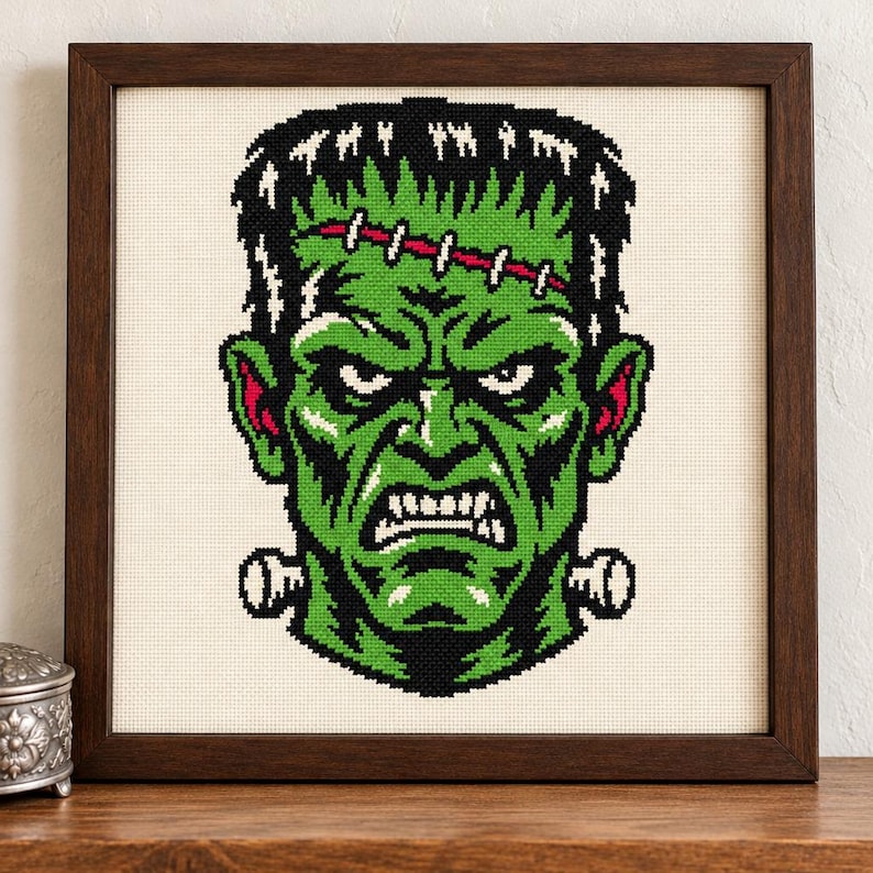 Puede incluir: Un bordado en punto de cruz enmarcado que muestra un monstruo de piel verde con cabello negro, una cicatriz cosida en la frente y pernos met&aacute;licos en el cuello. El dise&ntilde;o utiliza hilo negro, blanco y rojo sobre tela color crema en un marco de madera.