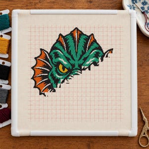 May include: A cross-stitch project on a square white frame featuring a partial creature design with green scales, orange fins, and a single yellow eye. The design is stitched onto a light beige grid fabric with red guide lines.