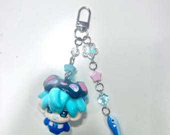 May include: A keychain featuring a small, stylized figure with bright blue hair and a dark hat, attached to a silver-tone metal clasp. A dangling chain includes a clear flower-shaped bead, a small pink star bead, a blue flower bead, and a blue fish charm.