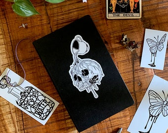 Op de afbeelding: Een zwart notitieboek met een witte sticker van een schedel waar een kopje vloeistof in giet. Het notitieboek ligt op een houten oppervlak met andere stickers, een tarotkaart en een kleine pompoen.
