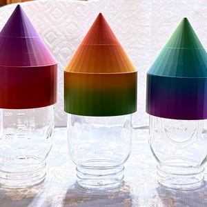 May include: Three clear glass mason jars with colorful, cone-shaped lids. The lids are in shades of purple, orange, and green. The jars have measurement markings on the side.