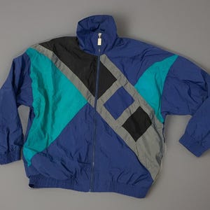 Peut inclure: Un coupe-vent à manches longues présentant un design color-block en bleu roi, sarcelle, noir et gris. La veste est dotée d'un col montant, d'une fermeture éclair frontale intégrale avec une tirette argentée, ainsi que de poignets et d'un ourlet élastiqués.