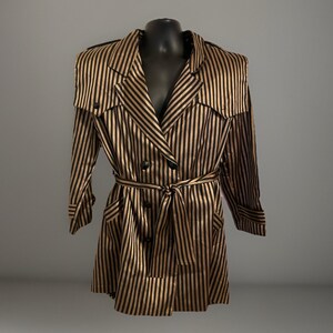May include: A double-breasted trench coat featuring a vertical striped pattern in metallic gold and black. The coat includes a notched lapel, long sleeves with cuffs, shoulder flaps, and a matching fabric tie belt at the waist with buttons.